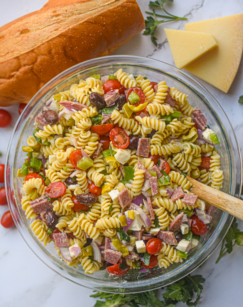 Italian pasta salad in a serving bowl with a wooden spoon surrounded by fresh bread and herbs