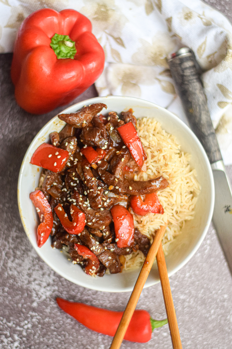Beijing Beef in a white bowl surrounded by chop sticks, red pepper and red chili pepper