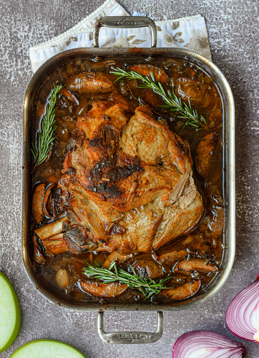 apple cider braised pork shoulder in a roasting pan surrounded by fresh thyme, onions, and apples