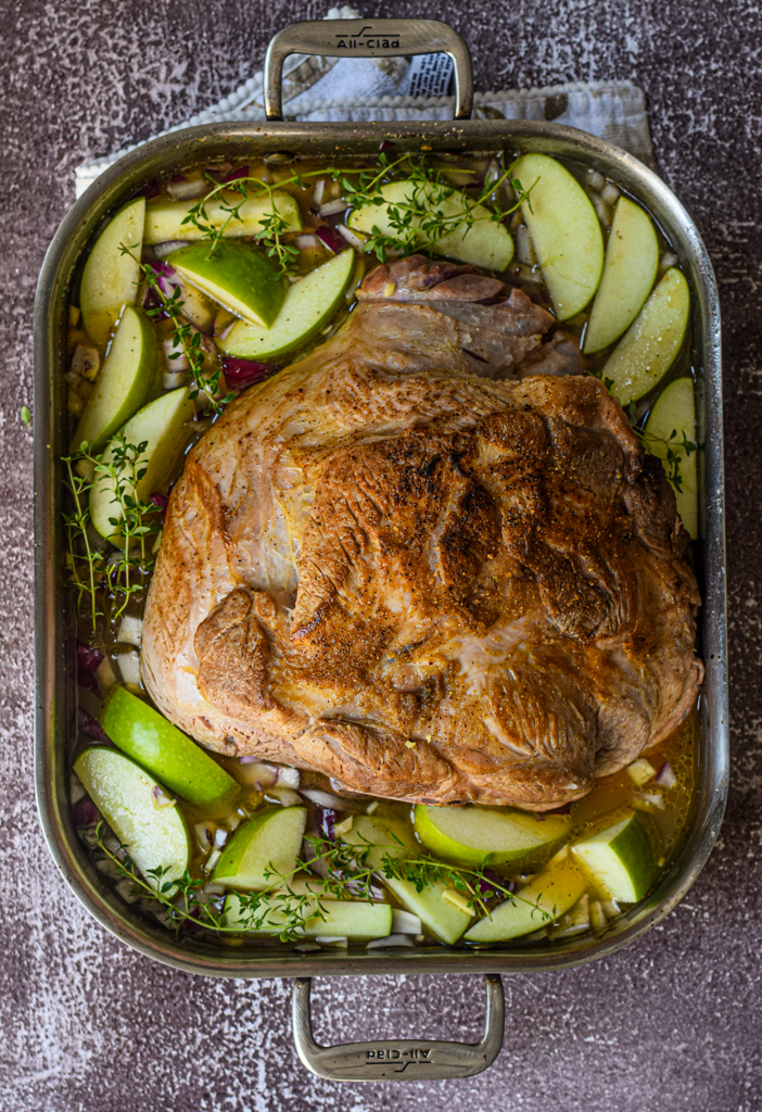 seared pork shoulder in a roasting pan surrounded by apples and onions in apple cider broth