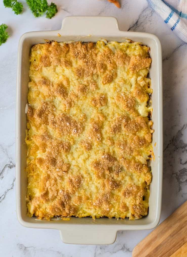 summer squash casserole in a white casserole dish surround by fresh herbs and a wooden cutting board