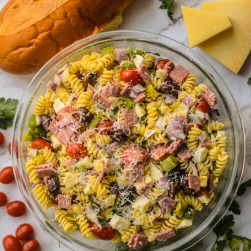 italian pasta salad in a glass bowl surrounded by fresh bread and herbs