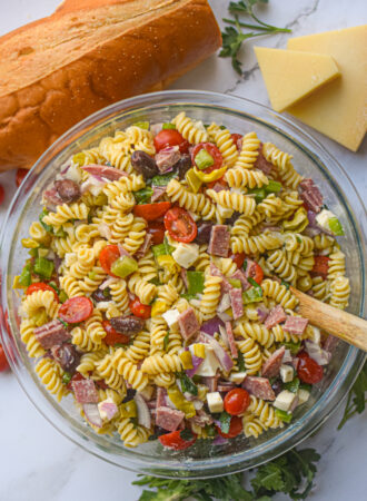 Italian pasta salad in a serving bowl with a wooden spoon surrounded by fresh bread and herbs