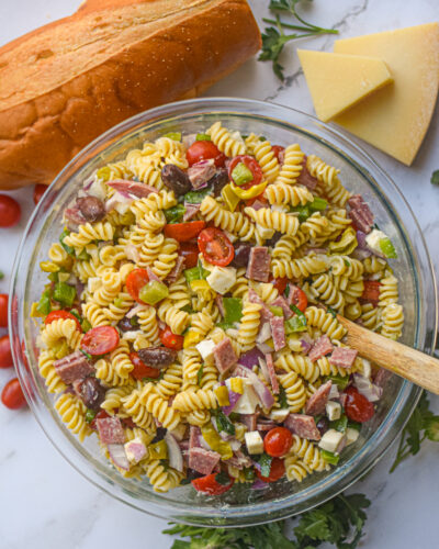 Italian pasta salad in a serving bowl with a wooden spoon surrounded by fresh bread and herbs