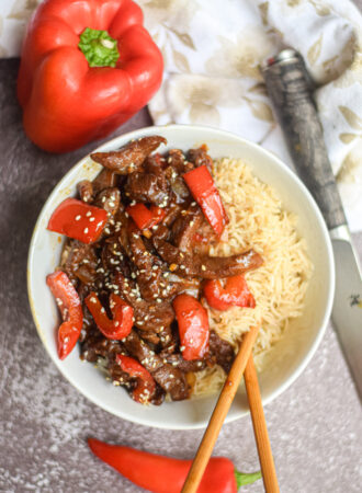 Beijing Beef in a white bowl surrounded by chop sticks, red pepper and red chili pepper