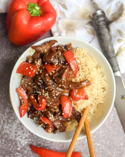 Beijing Beef in a white bowl surrounded by chop sticks, red pepper and red chili pepper