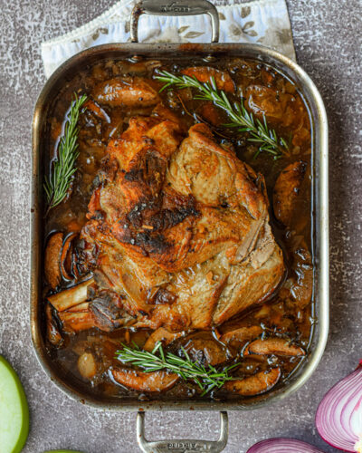 apple cider braised pork shoulder in a roasting pan surrounded by fresh thyme, onions, and apples